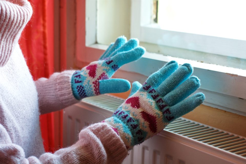 Person with gloves and winter clothes warms hands over a radiator