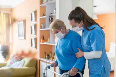 Home health aide wearing a facemask while assisting a senior woman walking with a mobility walker- personal care services