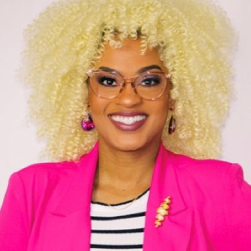 Woman with glasses and blonde curly hair smiles at the camera. She is wearing a bright pink blazer and a black and white striped shirt.