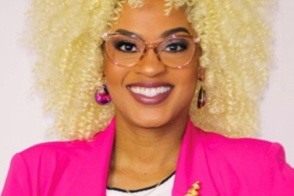 Woman with glasses and blonde curly hair smiles at the camera. She is wearing a bright pink blazer and a black and white striped shirt.