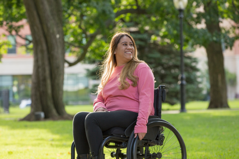Person smiling in a pink sweatshirt and dark pants, sitting in a wheelchair outside in the sun