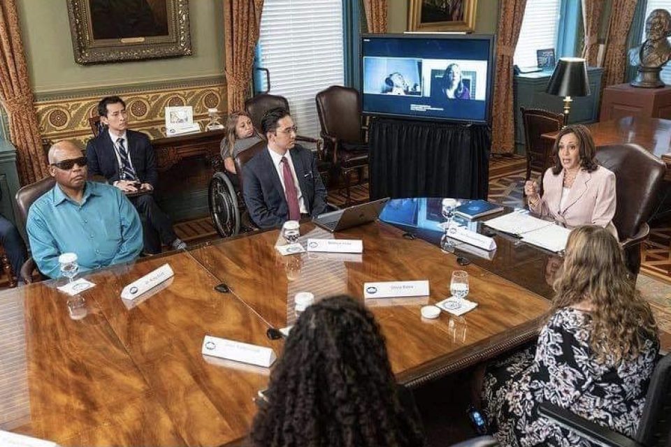 7 disabled voters meet with vice president Kamala Harris around conference table in DC. Two participants are participating virtually. Dr. Ricky Scott sits to the vice president's right. He is a black middle-aged man wearing a blue button down shirt and dark glasses. He has a serious expression on his face. The vice president is wearing a blush colored suit jacket and is speaking to the group, her right hand slightly raised above the table.. Three women, one in a wheelchair, and two men wearing suits are also in the room.