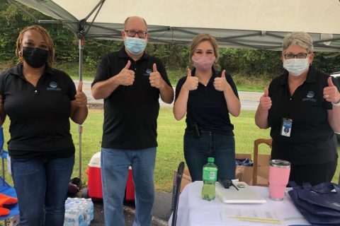 Four staff members of the Piedmont Triad Regional Council who partnered with DRNC on this event stand next to their booth, all wearing black polos and blue jeans, smiling from behind their masks. They all have two thumbs up. From left to right, a Black woman, older white man, a blond-haired woman and a white middle-aged woman.