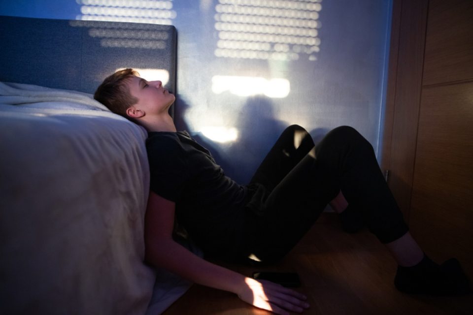A light-skinned boy with short blond hair sits on the floor of a darkened bed room in a prtf, light from the window reflects on the wall and his face.