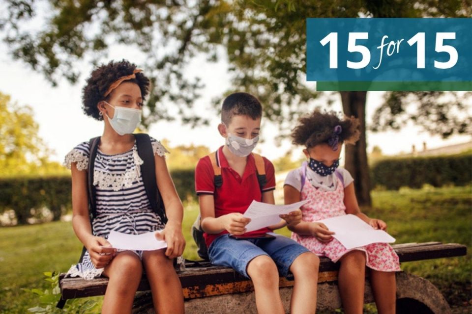 Three school aged kids with backpacks sitting on a park bench looking at school papers, while wearing medical masks
