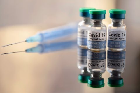 3 single use vials of COVID-19 Vaccine and on a reflective surface, a syringe lays nearby