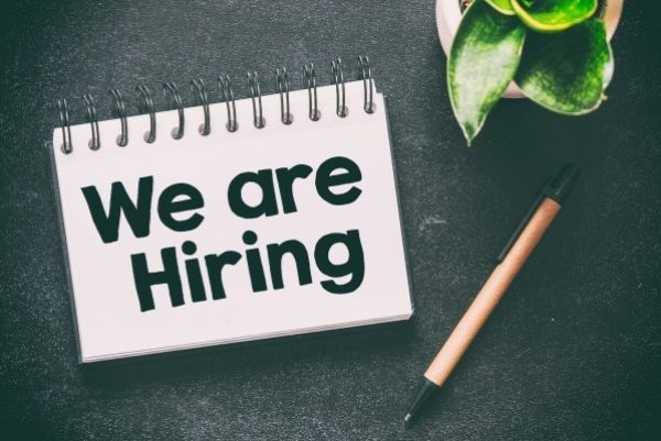 note pad with the words "we are hiring" on a table. a pencil and small plant lay to the side