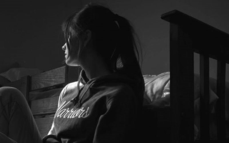 profile of a girl sitting on the floor by her bed in a prtf. She looks towards a window, her face obscured by her hair. She is sad.