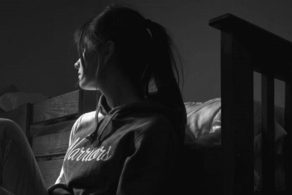 profile of a girl sitting on the floor by her bed in a prtf. She looks towards a window, her face obscured by her hair. She is sad.