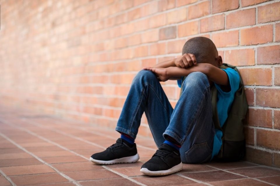 A young black boy is sitting by a long brick wall. His arms are crossed over his kness and his face his hidden.