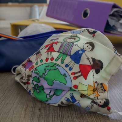 A white book bag with colorful print of the world and children from around the world holding hands and encircling the world. The bag is on a table with other back to school materials. A pencil case with a pencil sticking out and a purple binder full of paper are on the table behind the bag.