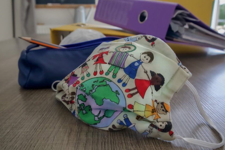 A white book bag with colorful print of the world and children from around the world holding hands and encircling the world. The bag is on a table with other back to school materials. A pencil case with a pencil sticking out and a purple binder full of paper are on the table behind the bag.