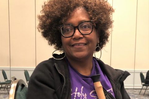Barbara Lee is smiling at the camera. She is wearing a purple t-shirt and black jacket. She is wearing black framed glasses and holding a cane.