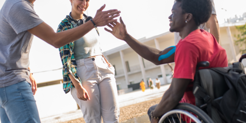 Three men and a woman are gaherred on the beach. They are smiling and look like they are enjoying themselves. A man in a wheelchair and another man are greeting each other.