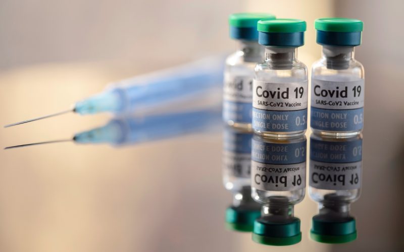 3 single use vials of COVID-19 Vaccine and on a reflective surface, a syringe lays nearby