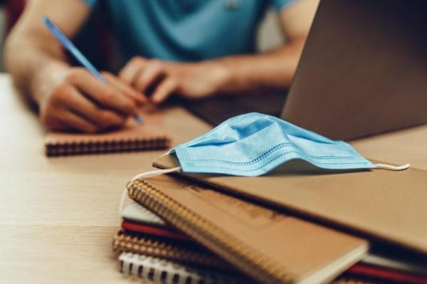 Person out of focus, completing an application, with a stack of books in focus and a COVID-19 face mask