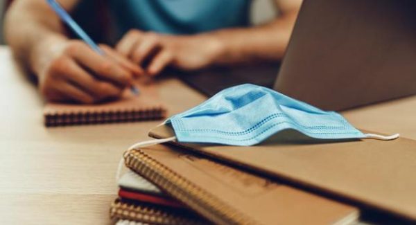 Person out of focus, completing an application, with a stack of books in focus and a COVID-19 face mask