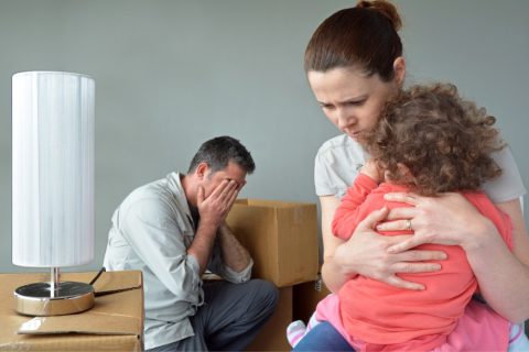 Evicted white family, father buries face in hands while mother holds toddler with a worried expression on her face. they are sitting in the middle of a bunch of moving boxes
