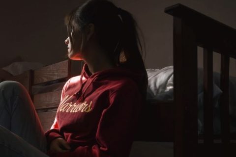 white girl with long black hair in pony wearing red sweatshirt sitting on the floor against her bed in a PRTF