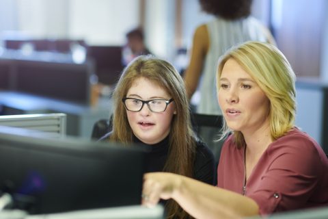 A young woman with an intellectual disability sits at a computer with her female boss, receiving training for her new job in the community..