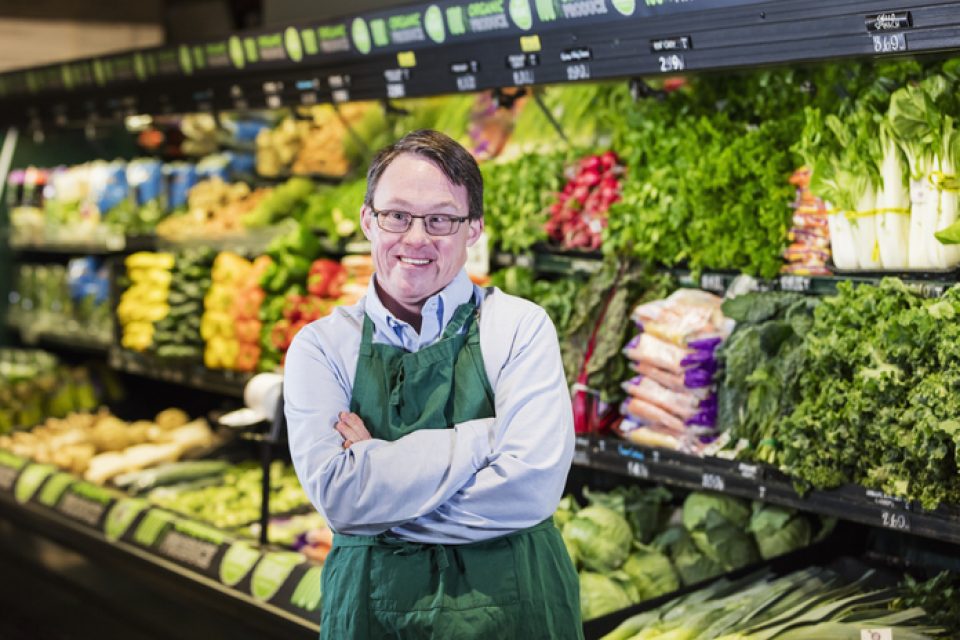a young man with glasses, brown hair and down syndrome wearing a grocery apron in a store where he works