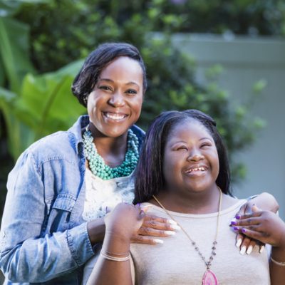 A woman with medium brown skin wearing a jean jacket and turquoise necklace smiles at the camera, her hands on her daughter's shoulders. Her daughter is wearing a bright pink pendant, a beige shirt and has a huge smile on her face and closed eyes. She has an intellectual disability.