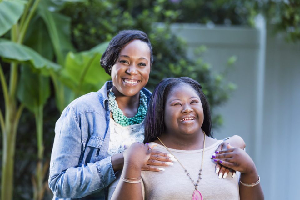 A woman with medium brown skin wearing a jean jacket and turquoise necklace smiles at the camera, her hands on her daughter's shoulders. Her daughter is wearing a bright pink pendant, a beige shirt and has a huge smile on her face and closed eyes. She has an intellectual disability.