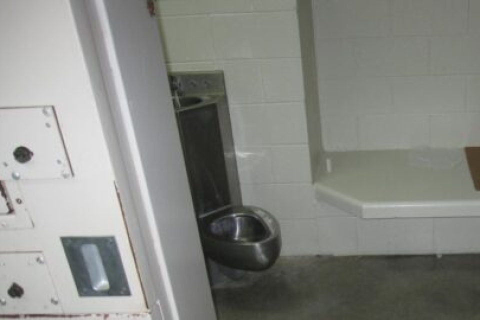 inside a solitary cell. a narrow tile shelf for a bed directly next to a steel toilet with a sink attached at the top