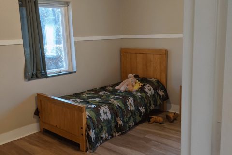 a child's bed in an empty room of an NC PRTF facility. worn stuffed animals on the bed and abandoned untied shoes on the floor. small window is over the bed.