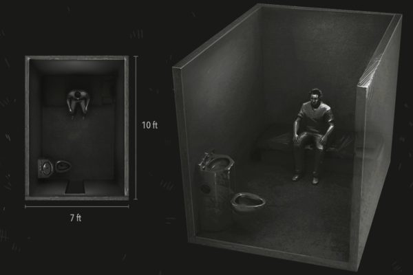 A very dark black and white overhead view of a man sitting alone in a solitary confinement cell.