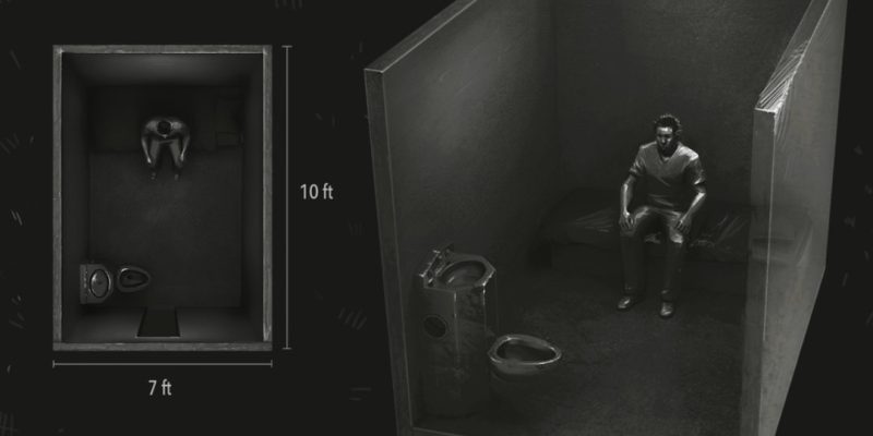 A very dark black and white overhead view of a man sitting alone in a solitary confinement cell.