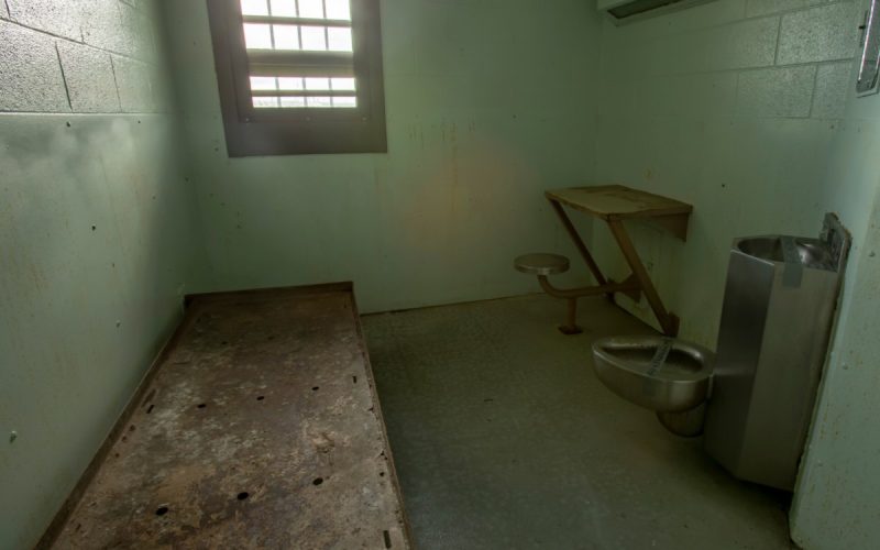 image of a solitary confinement cell - plank to lay on, small window, toilet that is also a sink, and small desk attached to concrete wall
