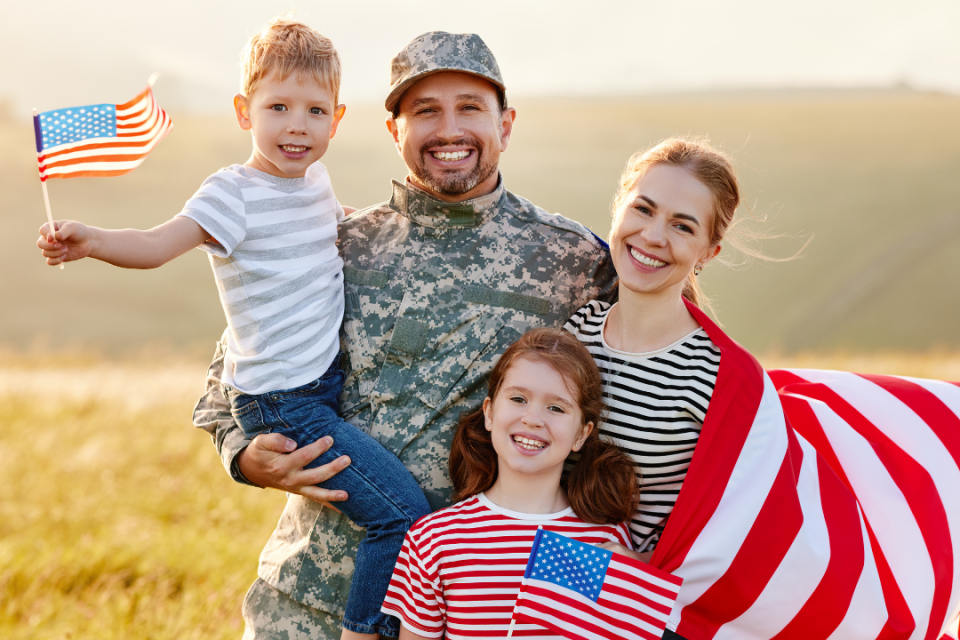 A man in uniform stands with his wife and two children. The are holding an american flag and smiling at the camera.