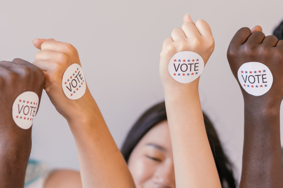 Three individuals putting their arms up with vote stickers on them.