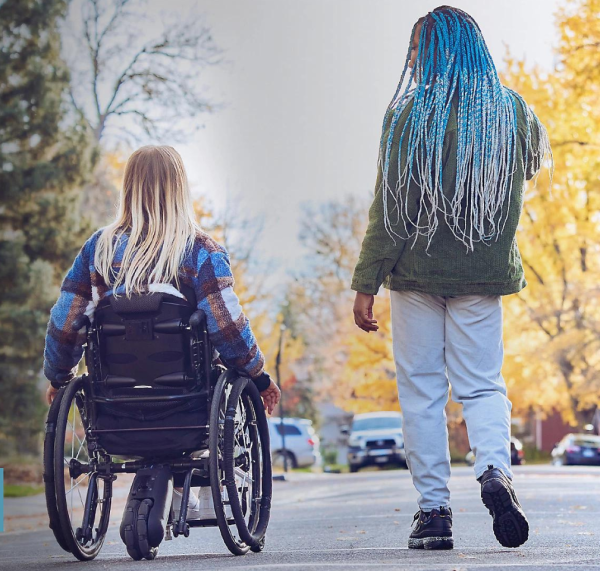 Two women are moving away from the camera. One is in a wheel chair and one is walking. They appear to be talking to each other.