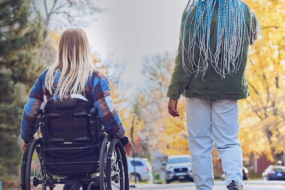 Two women are moving away from the camera. One is in a wheel chair and one is walking. They appear to be talking to each other.